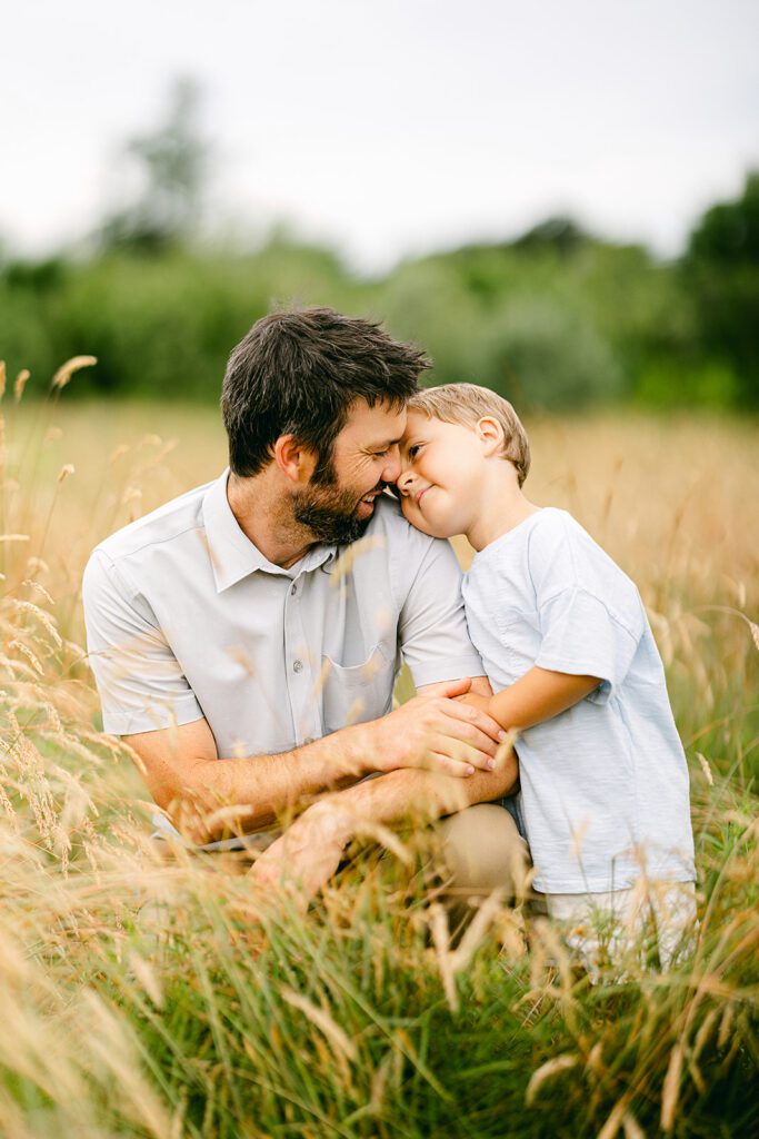 Dad squats in a field while his young son stands next to him and holds onto his arm; dad and son lean into each other with their heads touching.