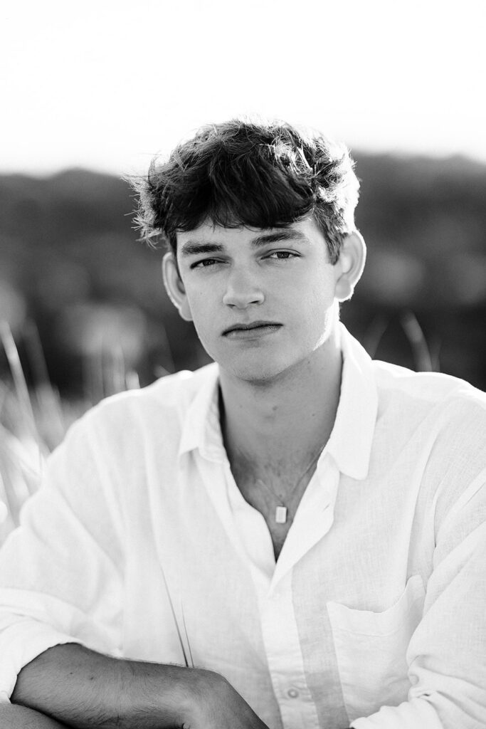 Black and white photo of a young man in a white linen shirt posing and look at the camera in front of a sand dune on Nantucket.