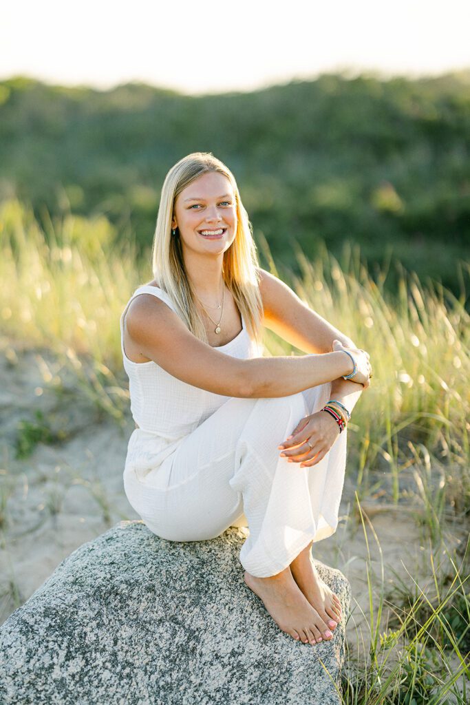 Young blond woman in a white matching set poses on a rock in front of a sand dune on Nantucket.