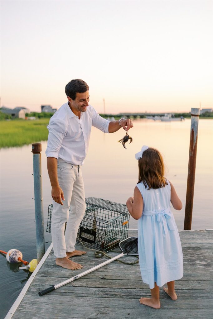 Dad and daughter standing on a dock in Madaket, Nantucket around sunset; Dad holds up a crab retrieved from the water.
