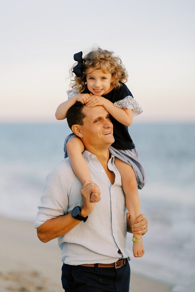 Dad holds young daughter with curly hair on his shoulders at the beach; she holds onto his head and smiles at the camera.