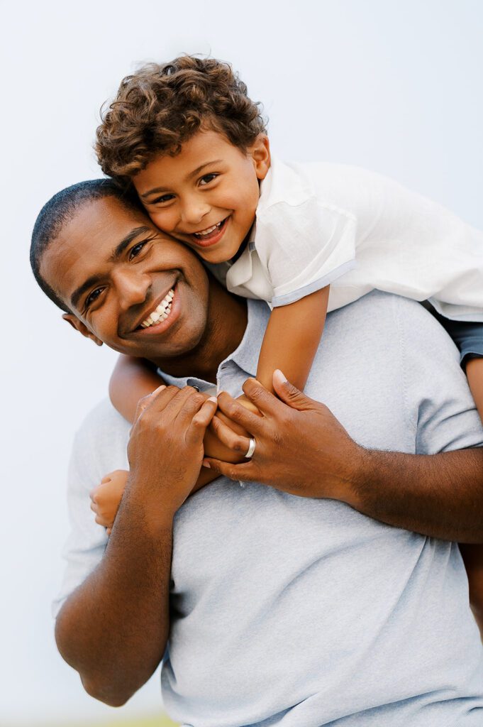 Boy on father's back with his arms wrapped around Dad's shoulders. Boy and Father both smile at camera.