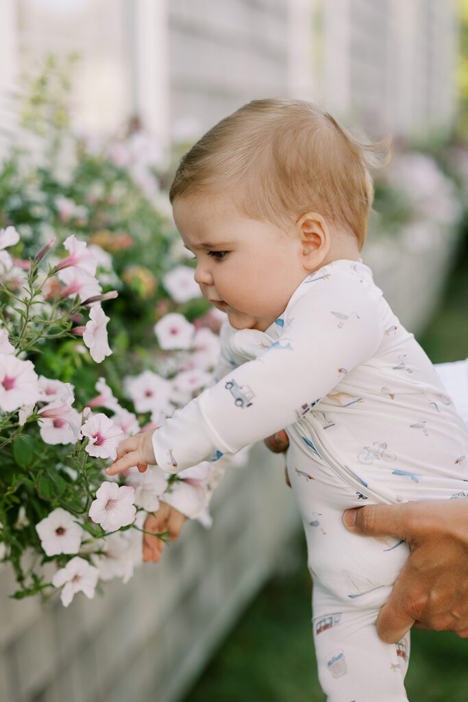 A sandy brown-haired baby in white pajamas with hand-drawn sketches featuring Nantucket imagery is reaching for some light pink flowers in a windowbox. 