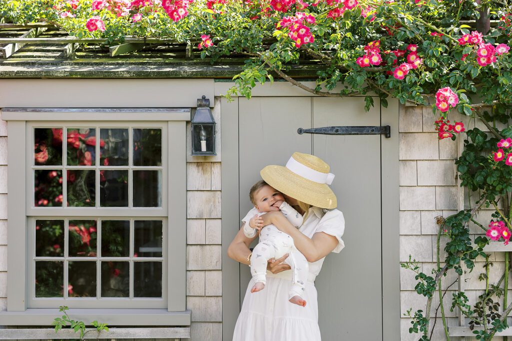 A mom wearing a white short sleeve dress and straw hat with white ribbon is holding her sandy-brown haired baby boy wearing pajamas with with hand-drawn sketches featuring Nantucket imagery and smooching his cheek while standing in front of a quintessential gray clapboard house with pink flowering vines above. 