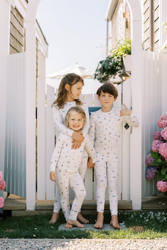 Three siblings are standing in front of the gate of a white picket fence wearing pajamas with hand-drawn sketches featuring Nantucket imagery. The older sister is standing with her arms draped around her younger sister who is smiling standing right in front of her, while their brother has a no-tooth smile and is holding a thumbs up. 