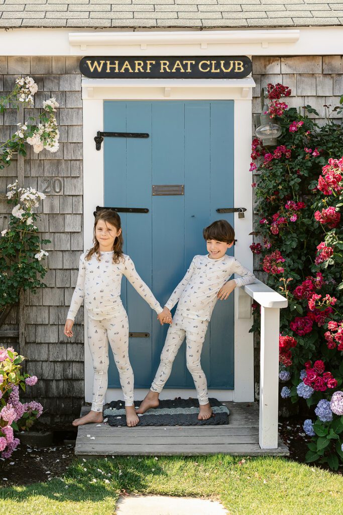 Brunette-haired boy/girl twins are  wearing pajamas with hand-drawn sketches featuring Nantucket imagery and holding hands while standing in front of the blue door of the Wharf Rat Club. 