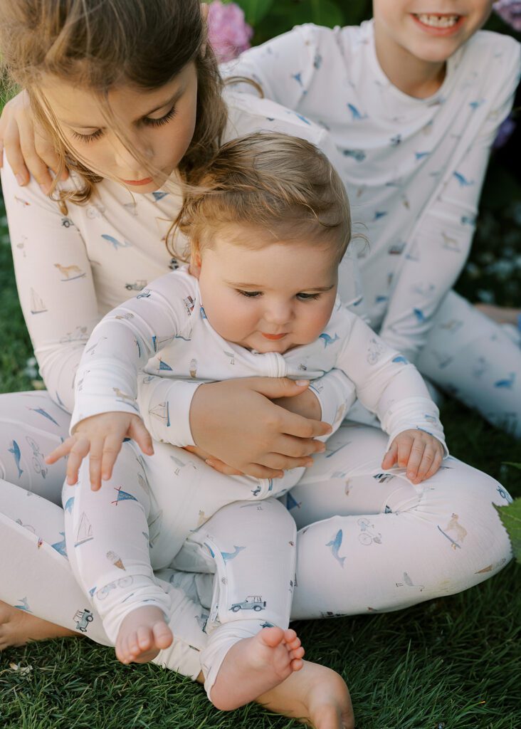 A young girl is sitting holding a chubby baby boy. Another child is sitting behind the girl and resting his hand on her shoulder - all three are wearing pajamas with hand-drawn sketches featuring Nantucket imagery.