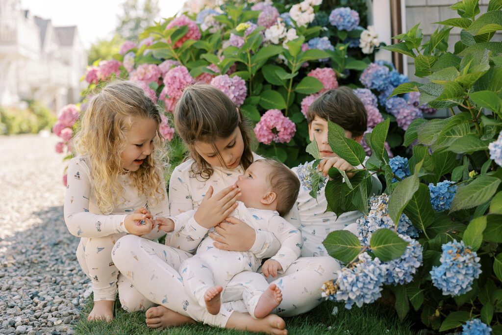 Four little kids are sitting in front of blooming hydrangea bushes wearing pajamas with hand-drawn sketches featuring Nantucket imagery. A brunette girl sitting in the middle is holding a baby boy. To her left is a sandy-brown haired little girl smiling over at the baby, and to her right is a brunette haired boy tucked behind a hydrangea branch. 