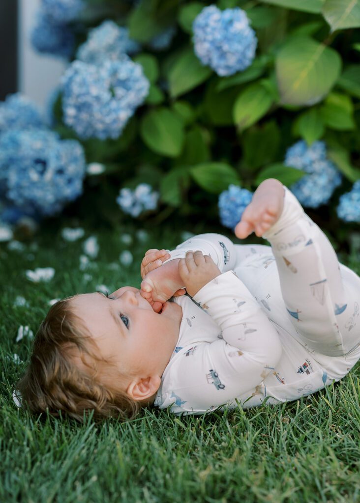 A sandy-brown haired baby boy  wearing pajamas with with hand-drawn sketches featuring Nantucket imagery is laying down on the lush green grass in front of a blue hydrangea bush putting his toes in his mouth. 