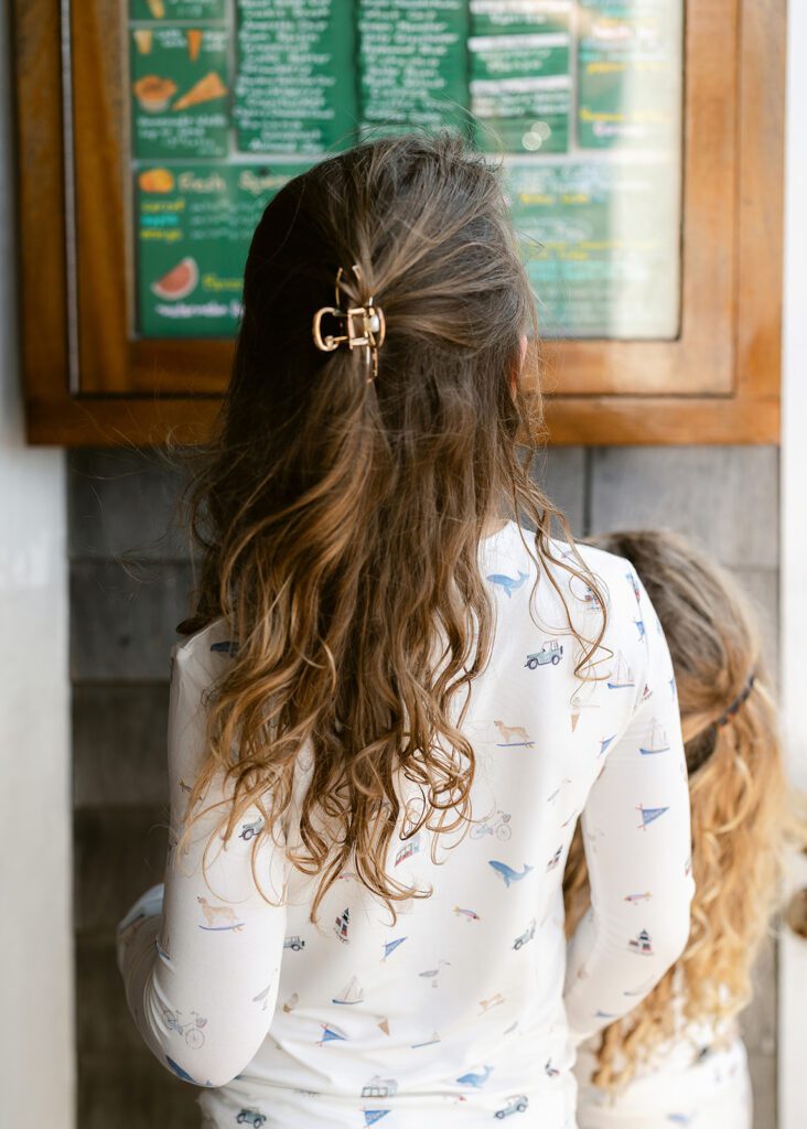A brunette girl is seen from behind with her beach curls hitting mid-back, wearing pajamas with hand-drawn sketches featuring Nantucket imagery and looking at the ice cream menu at The Juice Bar. 
