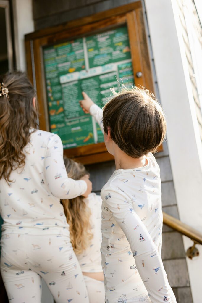 A brunette haired boy wearing pajamas with hand-drawn sketches featuring Nantucket imagery is pointing up at the menu outside The Juice Bar, while his two sisters in matching pajamas are standing in front of him. 