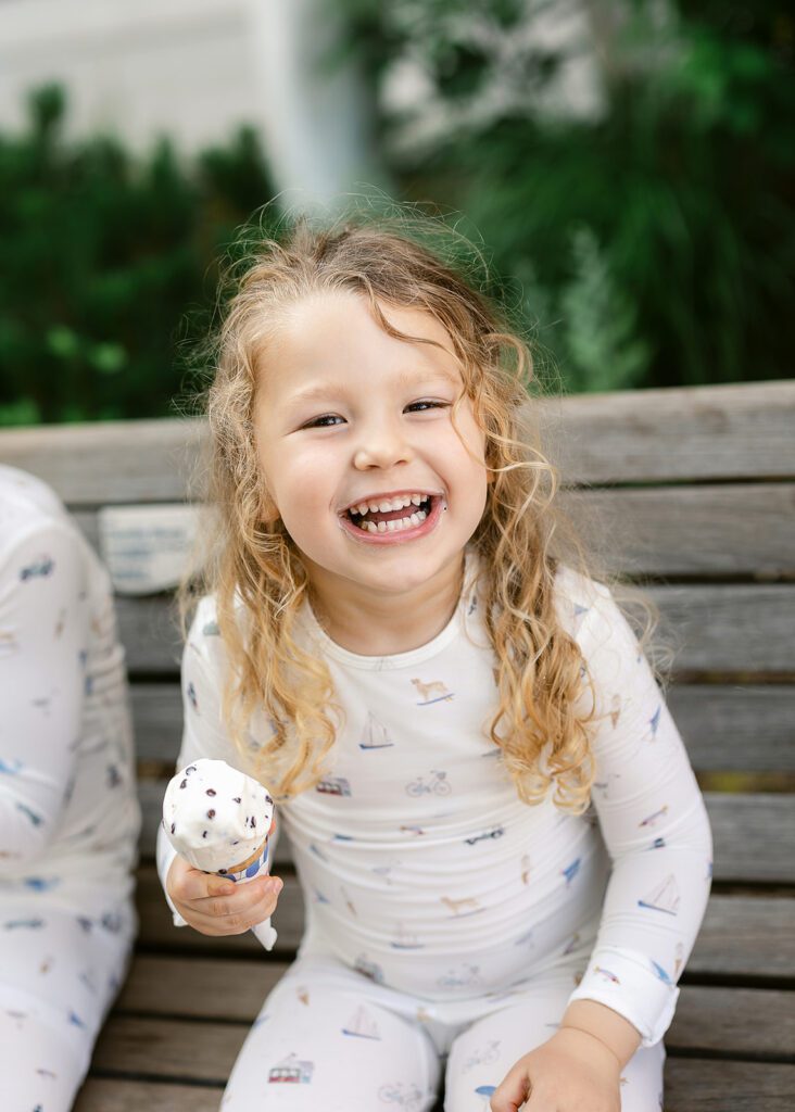 Curly-haired blond girl eating an ice cream cone on a bench, wearing printed pajamas.