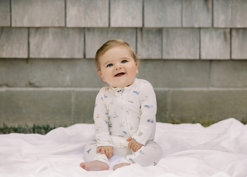 A sandy-brown haired baby boy  wearing pajamas with hand-drawn sketches featuring Nantucket imagery is sitting down on a white muslin blanket in front of a gray clapboard house and smiling. 