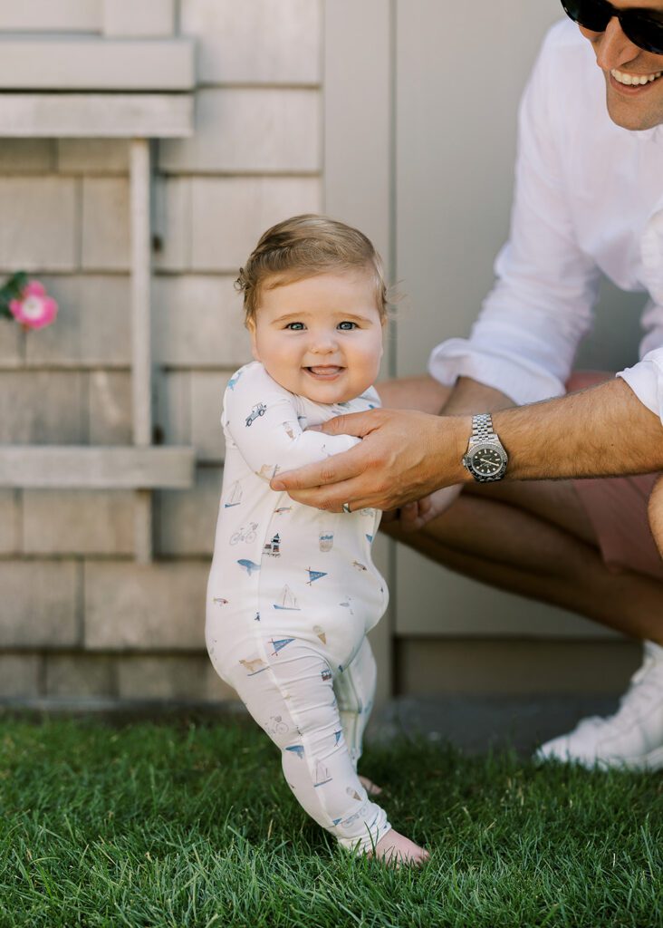 A baby is getting help from Dad standing on his wobbly legs atop lush green grass wearing pajamas with hand-drawn sketches featuring Nantucket imagery and smiling with his little tongue sticking out ever so slightly.