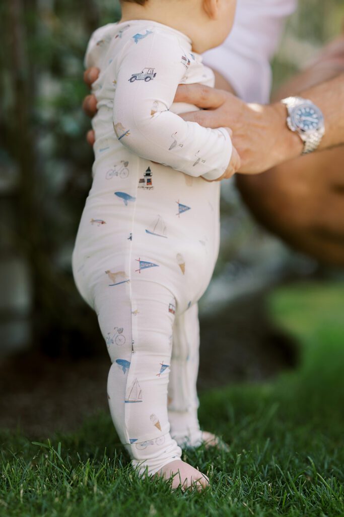 A baby is getting help standing on his wobbly legs on lush green grass wearing pajamas with hand-drawn sketches featuring Nantucket imagery such as bikes, pennants, whales, lighthouses, ice cream and more. 