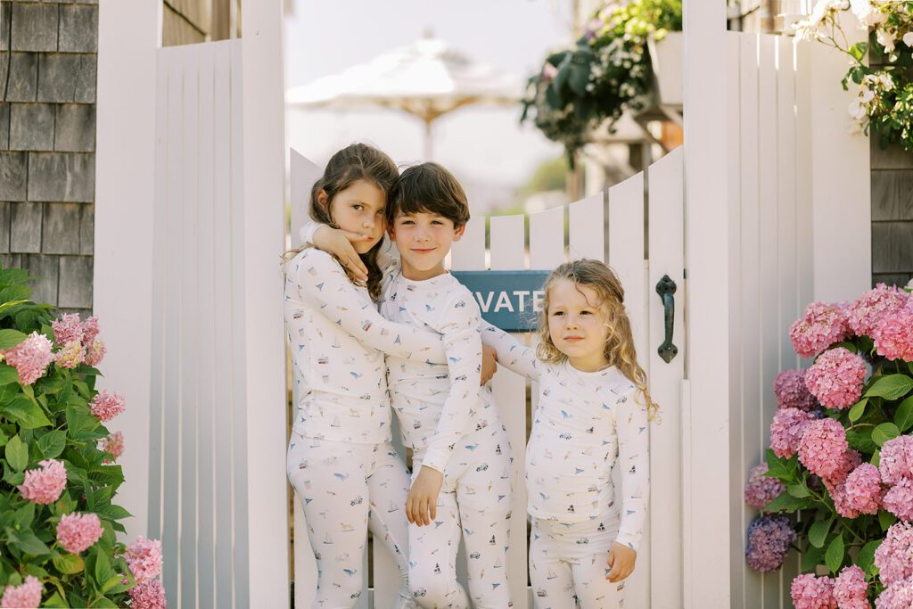 Three siblings are standing in front of a white picket fence door in matching pajamas with hand-drawn sketches featuring Nantucket imagery. The older two have dark brunette hair and have their arms around each other, while the youngest has sandy brown hair and has one hand on her brother's back. 
