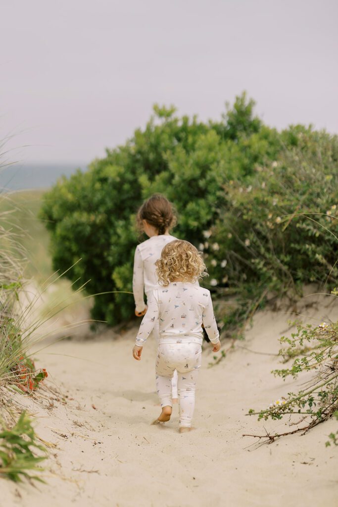 Two sisters, one blonde and one brunette, are wearing pajamas with hand-drawn sketches featuring Nantucket imagery and are walking along the sandy path in-between the grassy dunes leading towards the ocean.