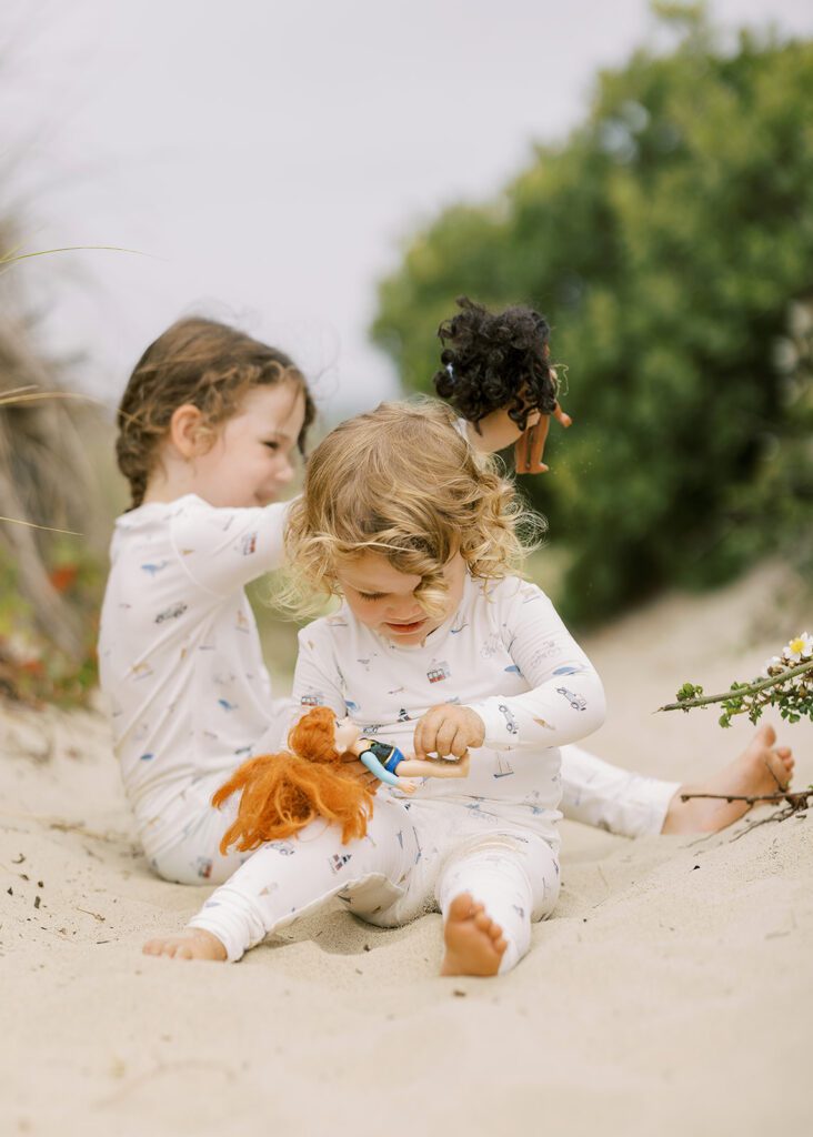 Two sisters, one blonde and one brunette, are wearing pajamas with hand-drawn sketches featuring Nantucket imagery, sitting in the same playing with their dolls. 