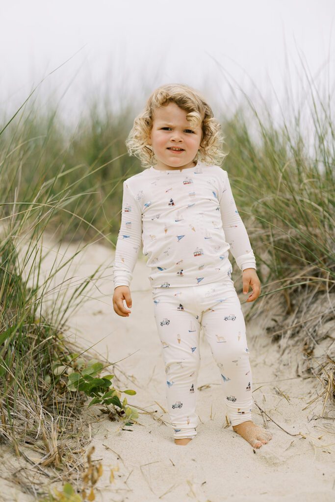 A blonde toddler girl wearing pajamas with hand-drawn sketches featuring Nantucket imagery is standing in sand surrounded by grassy dunes with a concerned look on her face and huge ringlet resting on her forehead. 