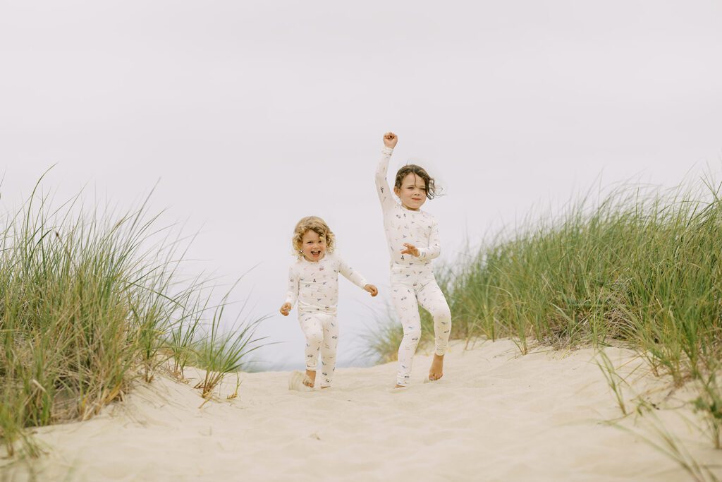 Two sisters, one blonde and one brunette, are wearing pajamas with hand-drawn sketches featuring Nantucket imagery are walking towards the ocean's edge. 