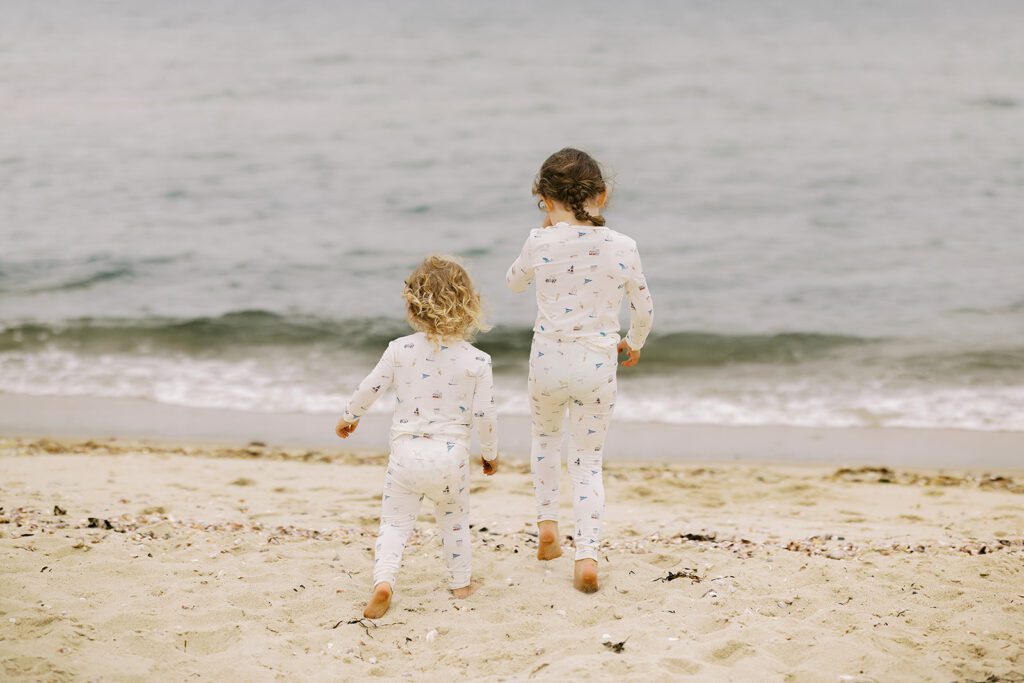 Two sisters, one blonde and one brunette, are wearing pajamas with hand-drawn sketches featuring Nantucket imagery and are walking towards the ocean's edge. 