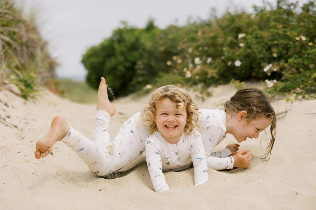 Two sisters are wearing pajamas with hand-drawn sketches featuring Nantucket imagery. The older brunette sister is lying on top of the younger blonde sister, perpendicular across her back, and both have huge smiles on their faces. 