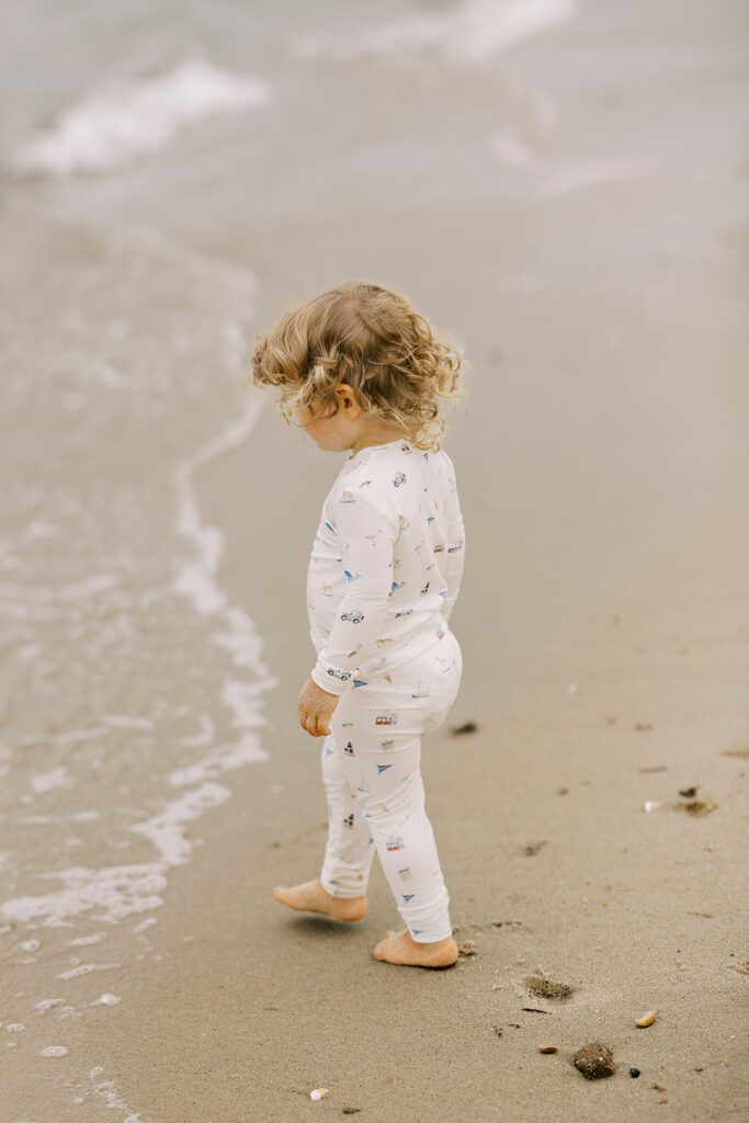 A blonde toddler girl wearing pajamas with hand-drawn sketches featuring Nantucket imagery is walking towards the ocean's edge. 