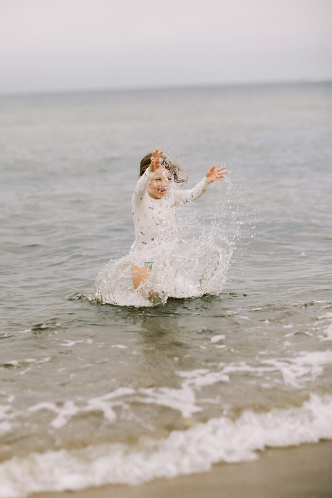 A brunette haired little girl is splashing in the ocean wearing wearing a pajama top with hand-drawn sketches featuring Nantucket imagery.