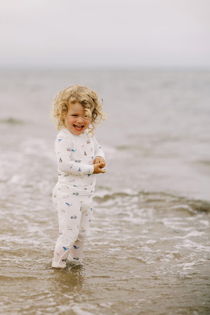 A blonde toddler girl wearing pajamas with hand-drawn sketches featuring Nantucket imagery has her feet covered by the ocean water with a huge smile on her face in delight! 
