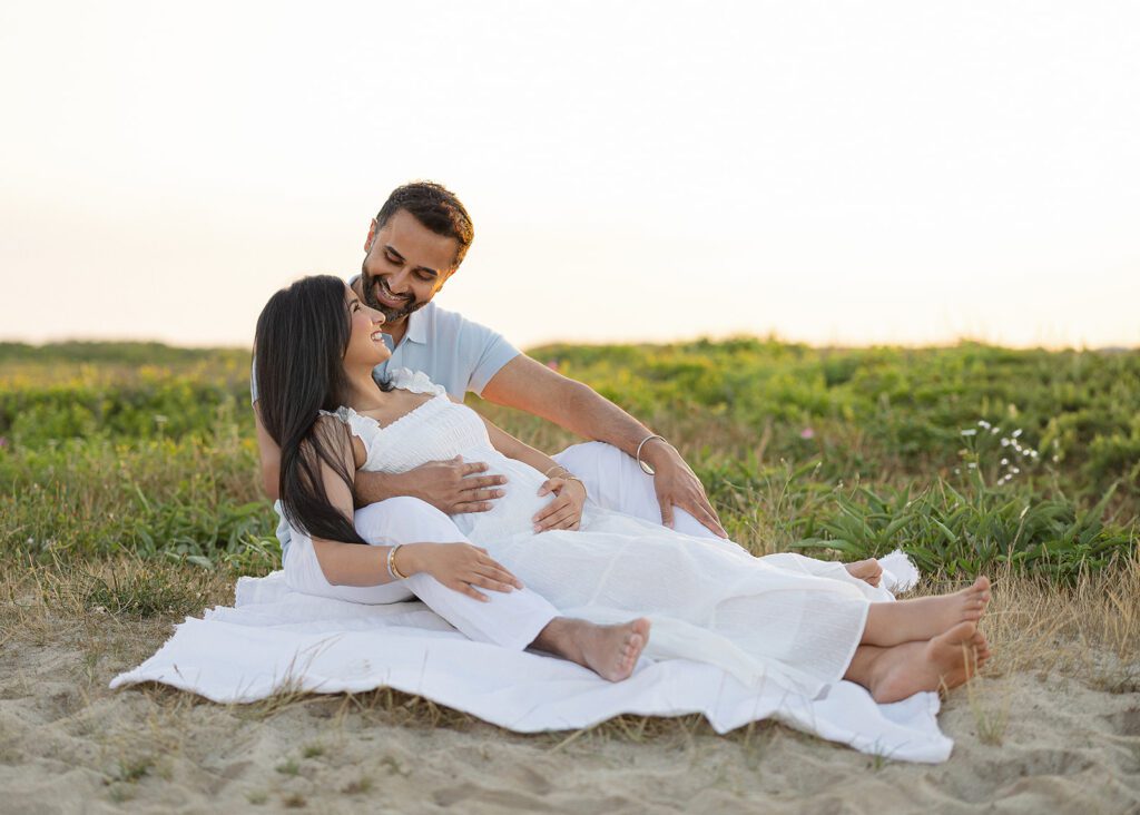 A man in a light gray shirt and white slacks is sitting behind his wife, a dark-haired woman in a white dress. She is sitting between his legs leaning back into him and smiling up at him. They are both barefoot and sitting on a white muslin blanket on the sandy, grassy dunes. 