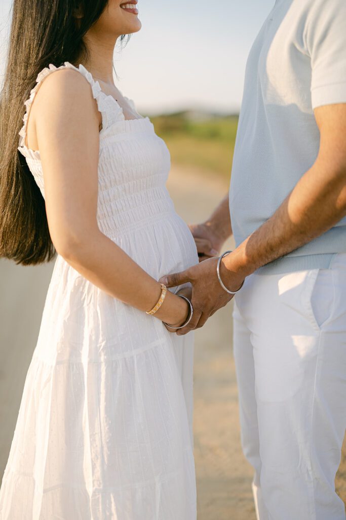 A dark-haired woman in a white dress is facing a man in a light gray shirt and white slacks - they are holding hands and you can see that she is smiling while his head is not in the photo. 