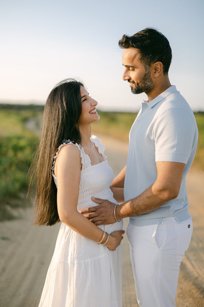 A dark-haired woman in a white dress is facing a man in a light gray shirt and white slacks. She is cupping her pregnant belly and smiling up at him, while he is smiling down at her and resting his hands on her belly/waist. 