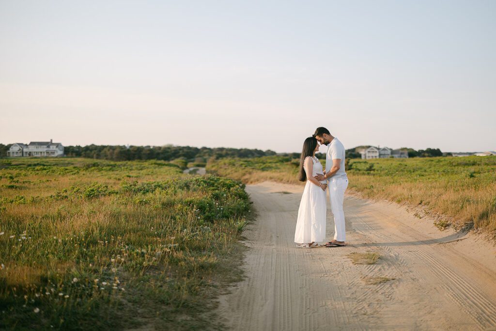A dark-haired man and woman are standing on a sandy drive between grassy fields facing each other with foreheads touching, all hands resting on her pregnant belly. 