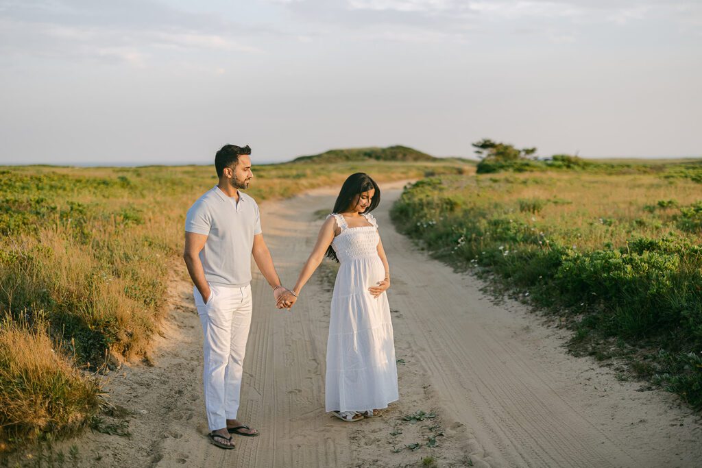 Dark-haired man in a gray colored shirt and white slacks is holding the hand of a dark-haired woman in a white dress who is cupping her pregnant belly with her other hand. They are standing on a sandy drive between two grassy fields and he is looking at her while she is looking down at her belly.