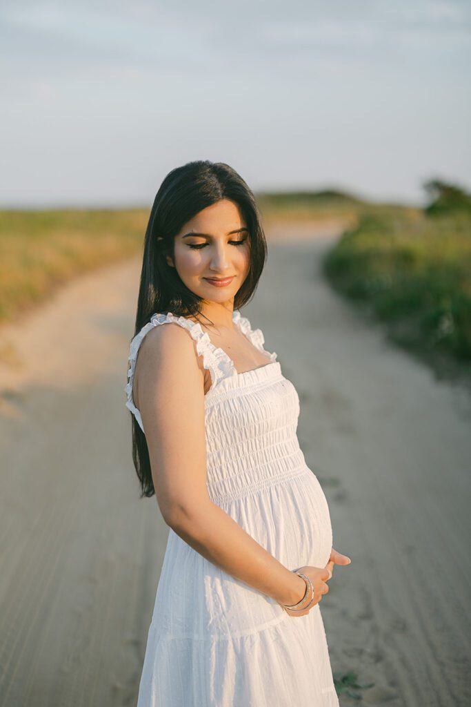 A woman in a white dress with dark hair cascading down her back is standing in a sandy lane cupping the bottom of her pregnant belly and looking over her shoulder down at the ground. 