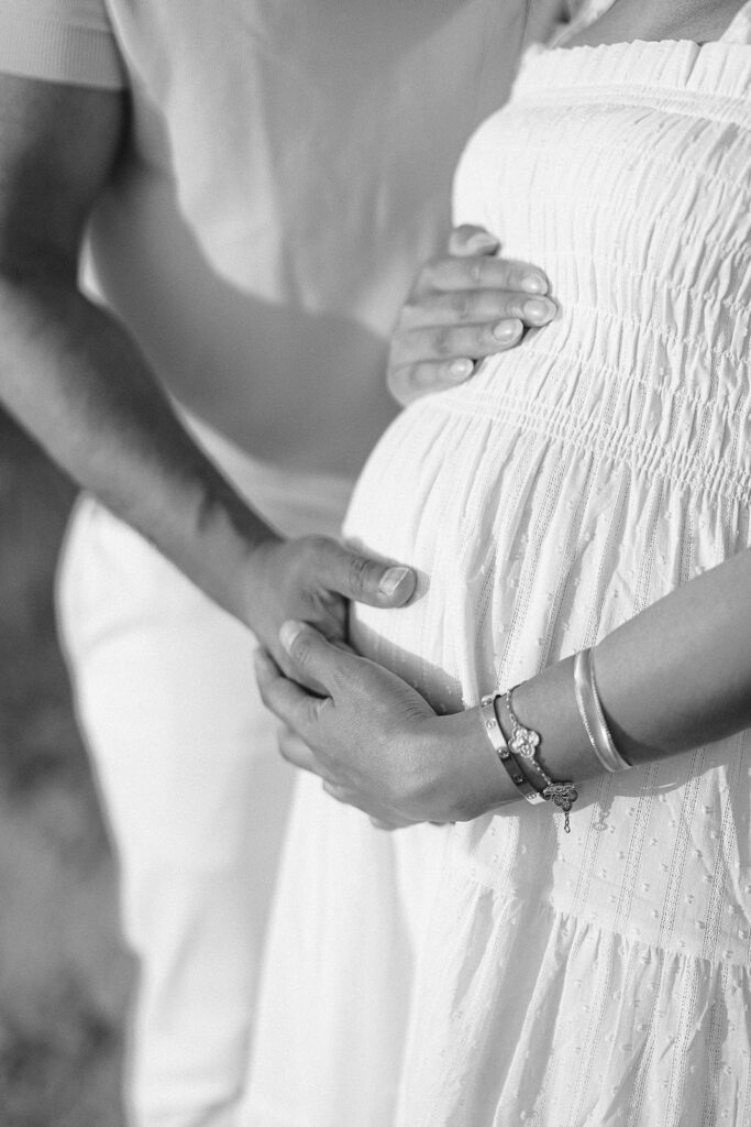 A pregnant woman in a dress is standing with her hands cupping her preganent belly. Her husband is standing beside her and he is also resting his hand on her belly. 