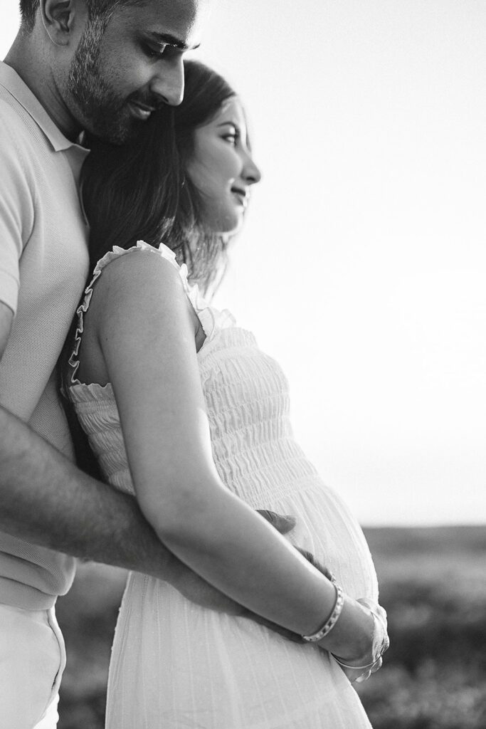 A man is standing behind his wife with his arms around her, hands resting on her pregnant belly. She is leaning back into him and smiling, hands cupping her belly. 