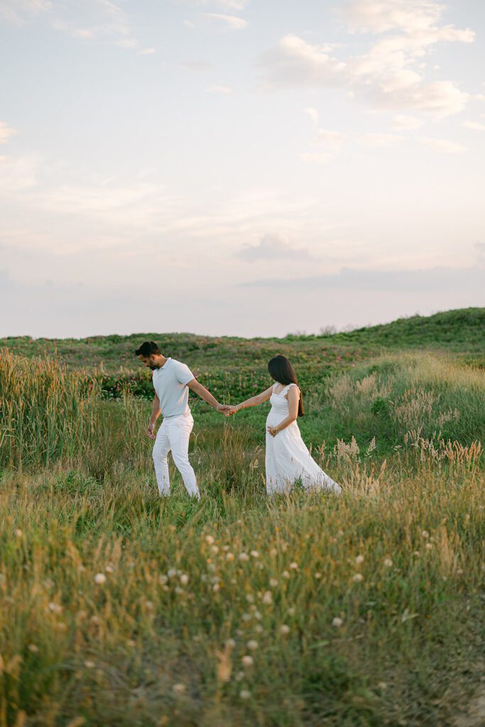 A dark-haired man wearing white slacks and a light gray shirt is holding his wife's hand, leading her through a grassy field. She has long dark hair and is wearing a long white dress, resting one hand on her pregnant belly. 
