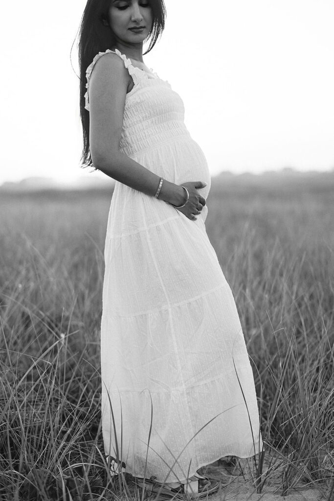 A woman in a long dress with dark hair cascading down her back is standing in a grassy field cupping the bottom of her pregnant belly and looking over her shoulder down at the ground. 