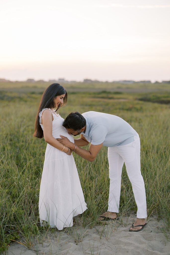 A dark-haired woman in a white dress is facing a man in a light gray shirt and white slacks. She is cupping her pregnant belly and smiling down at him, while he is bending over and kissing her pregnant belly. 