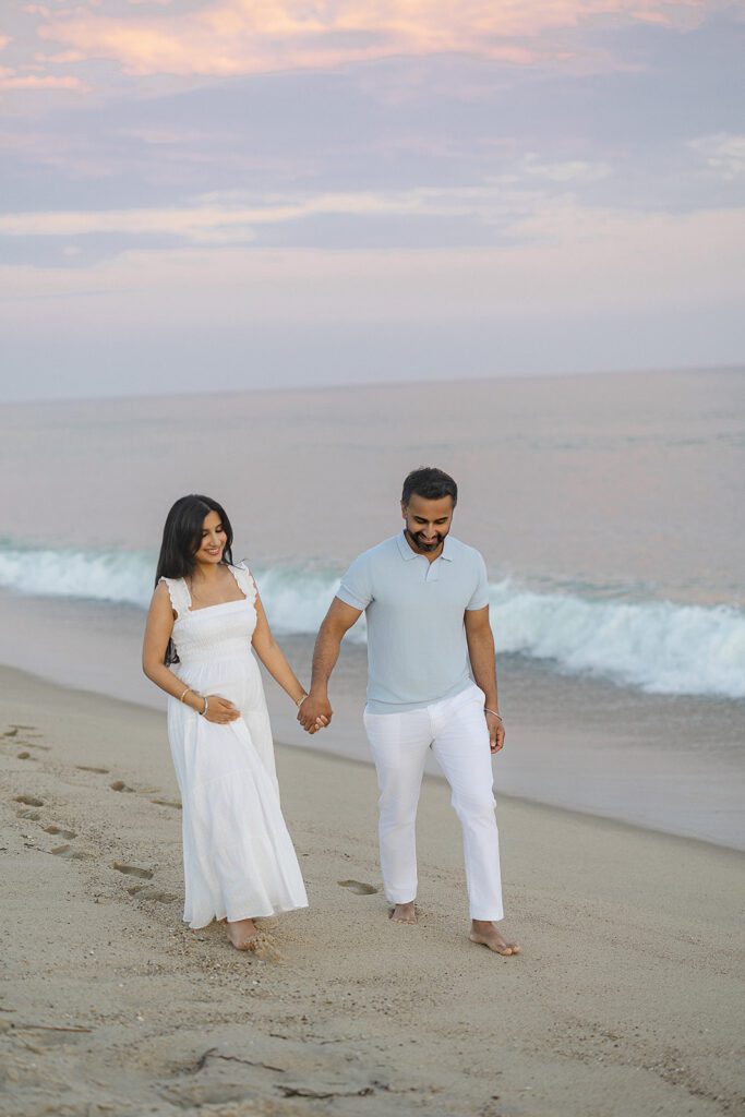A pregnant woman with long dark hair is barefoot wearing a long white dress, holding her husbands hand who is wearing white slacks and a light gray shirt. Both are smiling as they walk on the beach along the ocean's edge. 
