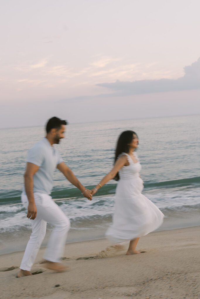 A dark-haired man and women are holding hands and walking along the waters edge at the beach. She is wearing a white dress and he is wearing white slacks and a light gray shirt.  