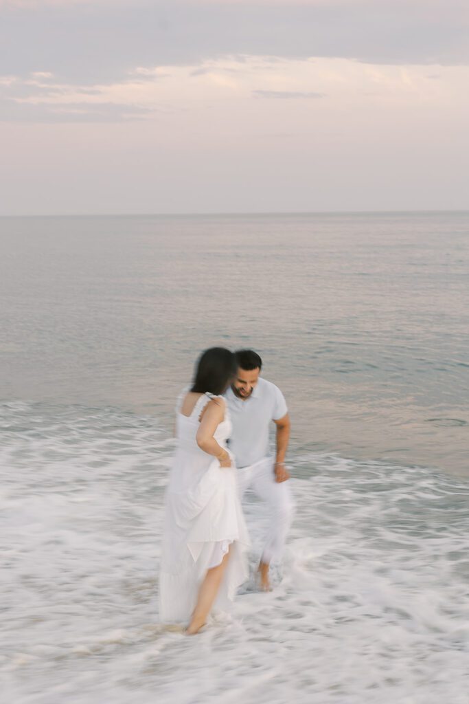 A dark-haired man and women are getting their feet wet in the ocean. She is wearing a white dress and he is wearing white slacks and a light gray shirt. 