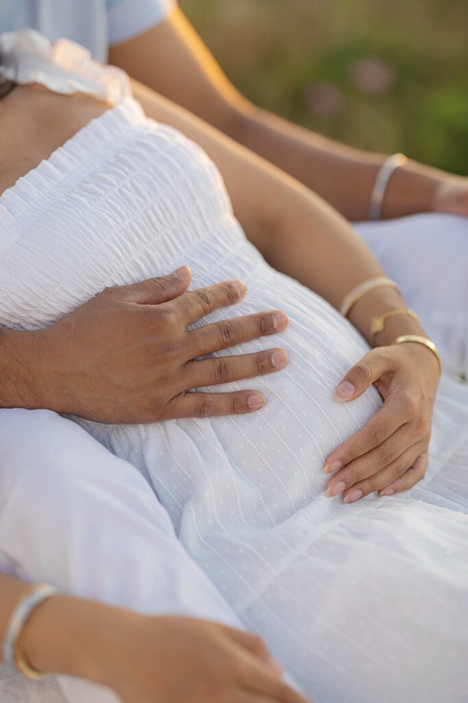 A pregnant woman is sitting between her husbands legs wearing a white swiss-dot cotton dress and gold bracelets, leaning back into him, each resting one hand on her belly. 