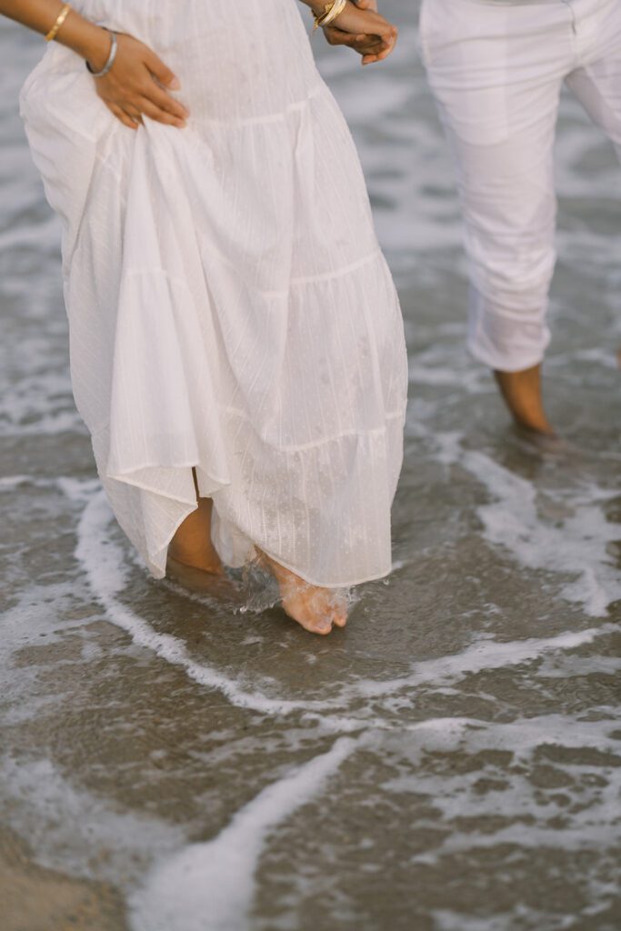 A woman in a long white dress and a man in white slacks are barefoot and walking in the ocean. 