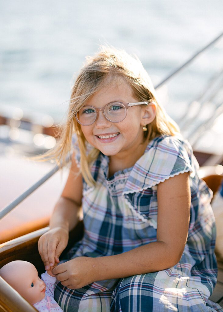 A young girl is smiling while sitting on the deck of the The Blue Peter Yacht sailboat on Nantucket, her baby doll dangling from her fingers. 