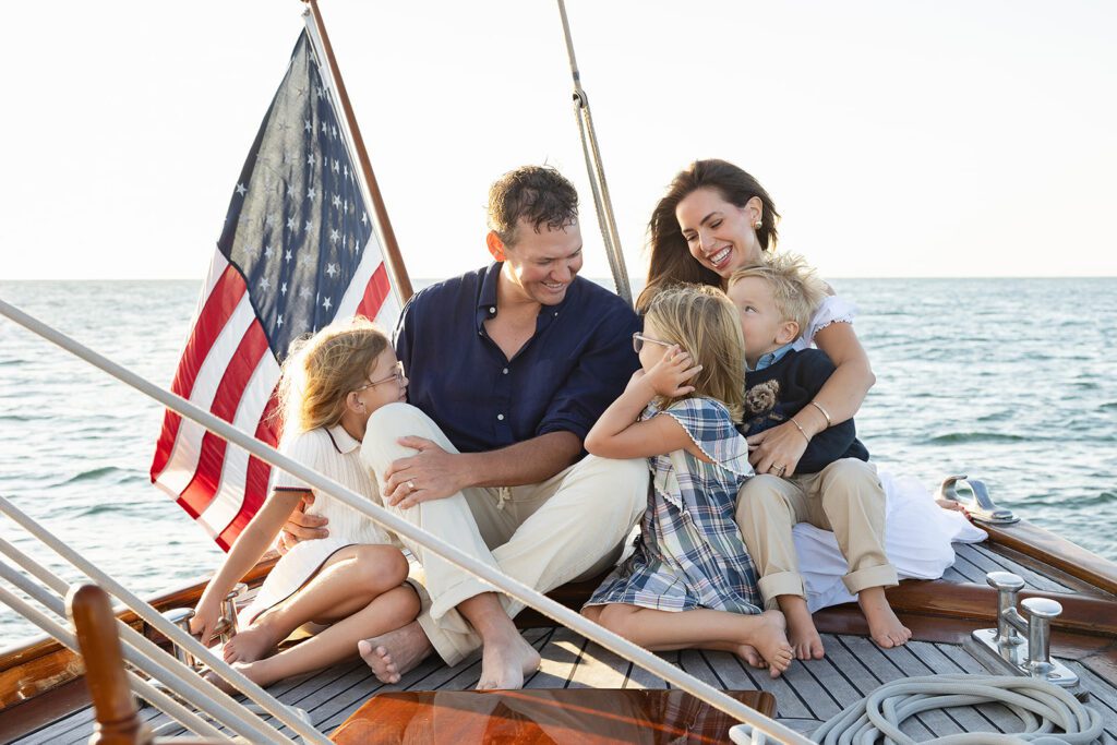 A family of five sits on the bow of The Blue Peter Yacht sailboat on Nantucket, smiling and laughing with one another. 