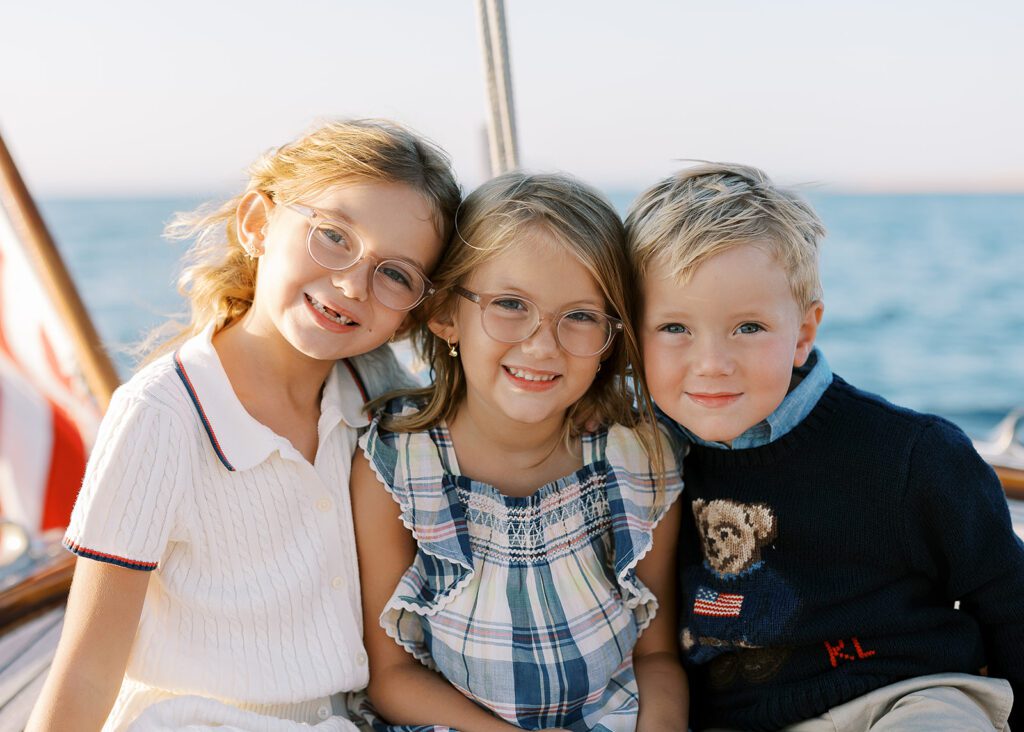 Three young siblings are sitting shoulder-to-shoulder on the deck of The Blue Peter Yacht sailboat on Nantucket and smiling. 