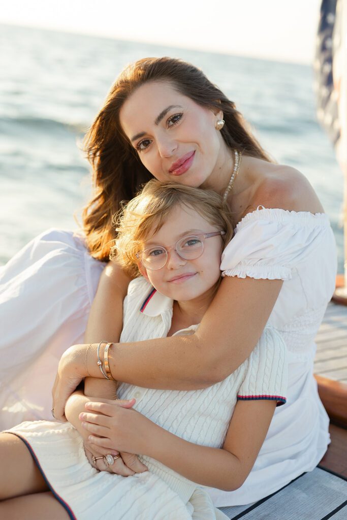 A mother and her daughter are sitting on the deck of The Blue Peter Yacht sailboat on Nantucket, embraced in a hug and smiling. 