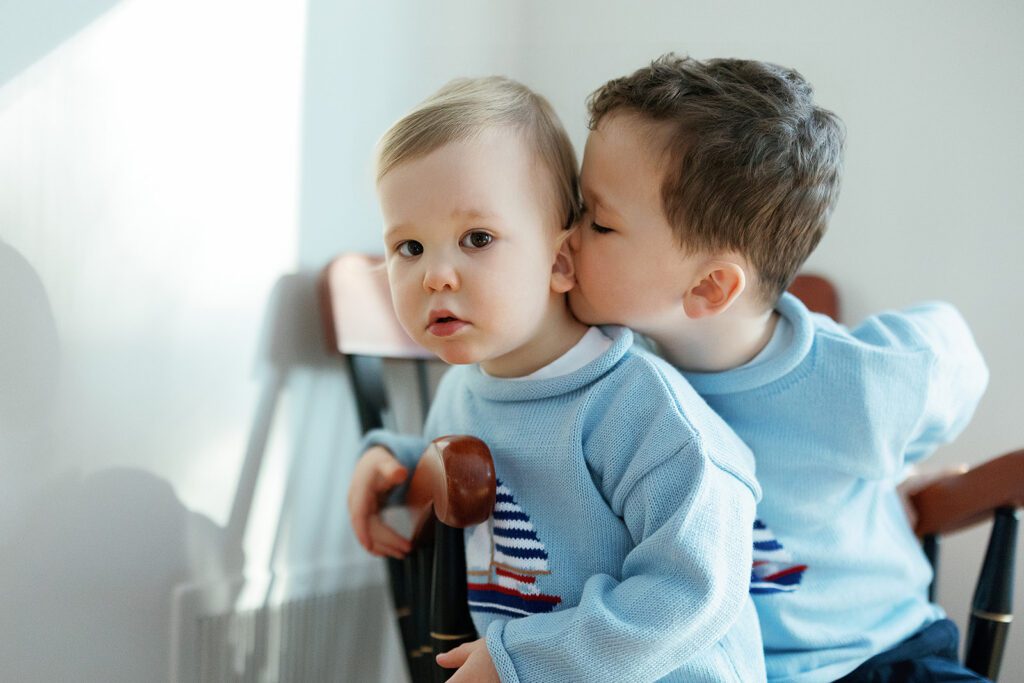 Two toddler brothers in matching sweaters are sitting together on a chair, the oldest planting a kiss on his little brother's neck at home in the Connecticut suburbs. 